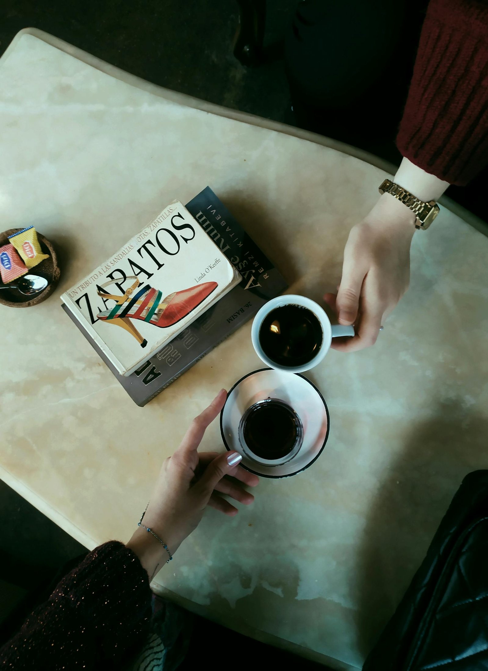 Two people enjoy coffee and discuss fashion book 'Zapatos' on a marble table.