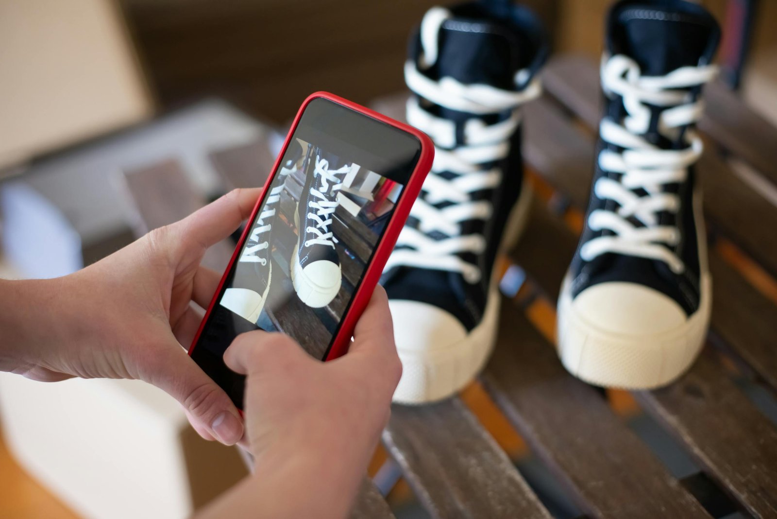 Hands using a smartphone to photograph black sneakers on a wooden surface.